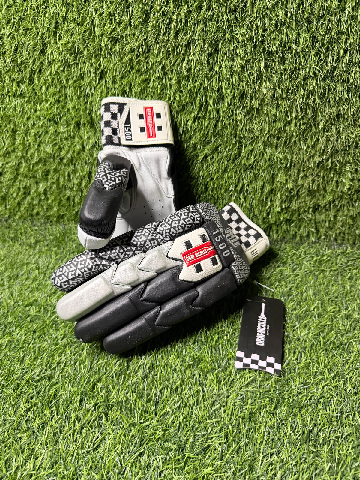 A pair of black and white racing gloves with geometric patterns and checkered details, displayed on green artificial turf with a brand tag attached.
