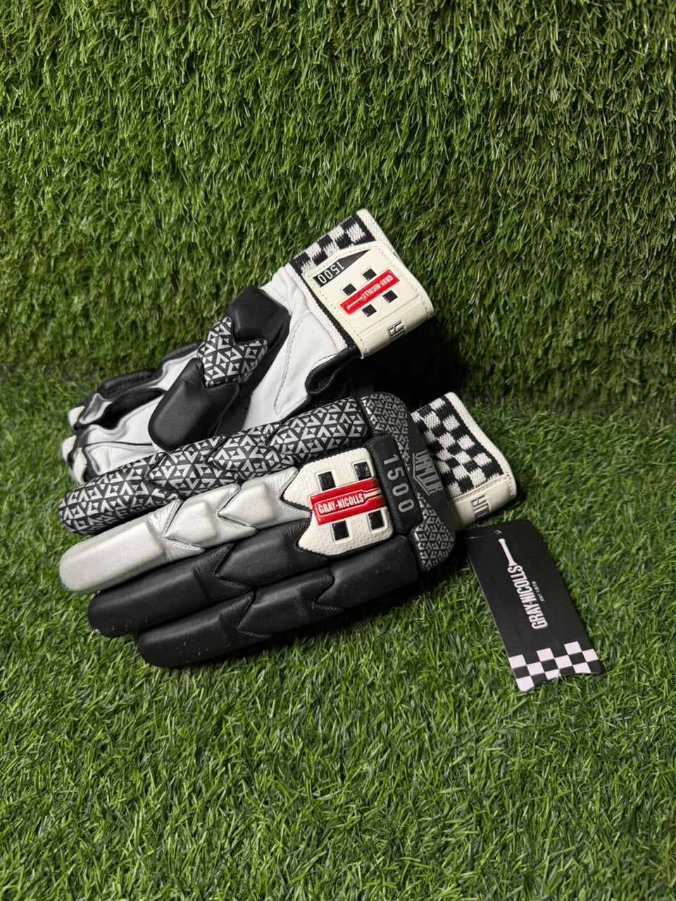 A pair of black and white racing gloves with geometric patterns and checkered details, displayed on green artificial turf with a brand tag attached.