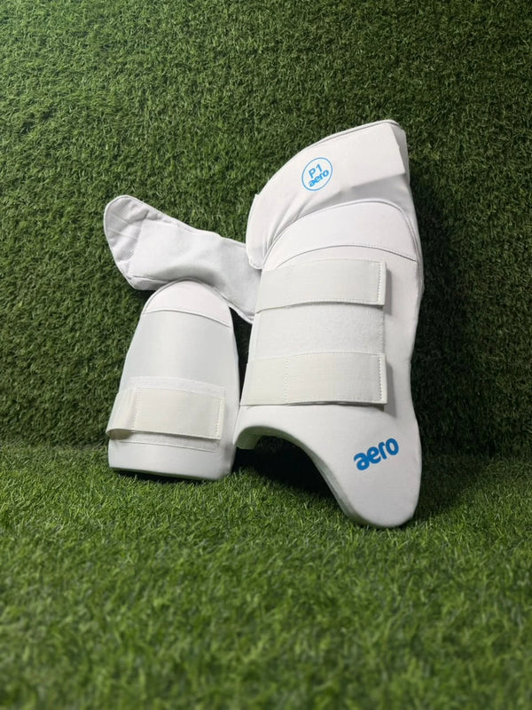 White Aero Baby inline skating shin guards with blue branding, displayed on a green artificial turf background.